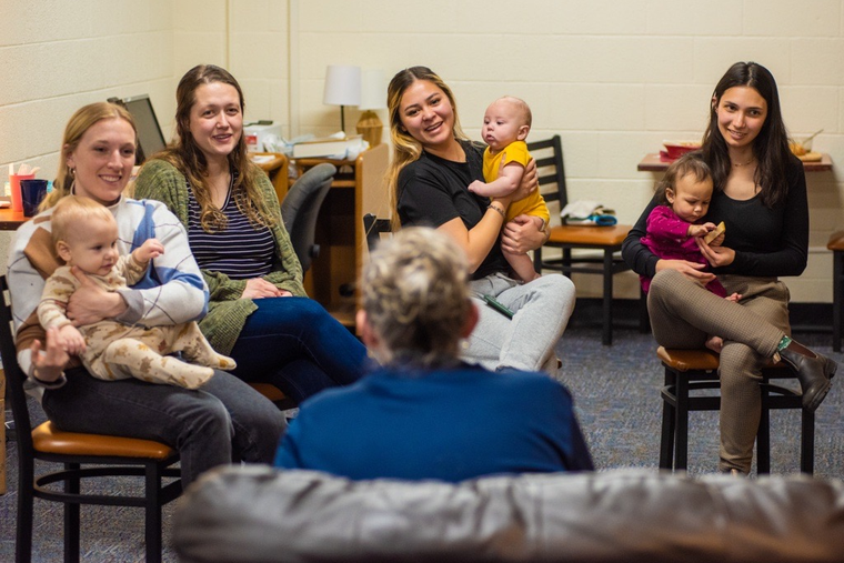 Young moms and students Angelina Hanft, shown center, and Katie, at right, next to each other with their babies, participate in a community night with university staff who are fellow moms at the University of Mary. 